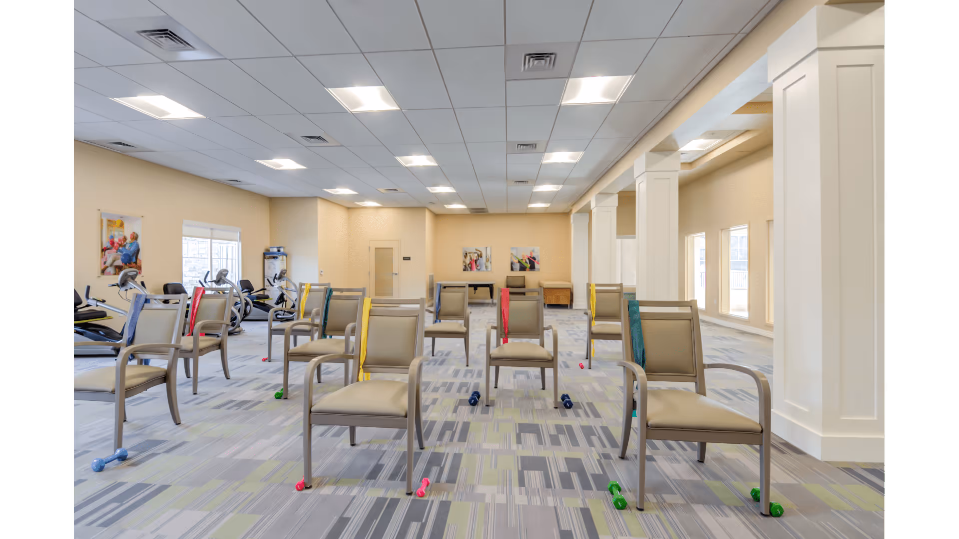 A spacious exercise room with multiple chairs arranged in rows, each chair paired with small colorful dumbbells on the floor. The room has large windows on the right side allowing natural light, light-colored walls, and a patterned carpet. Exercise equipment is visible in the back left corner, and there are framed pictures on the walls.