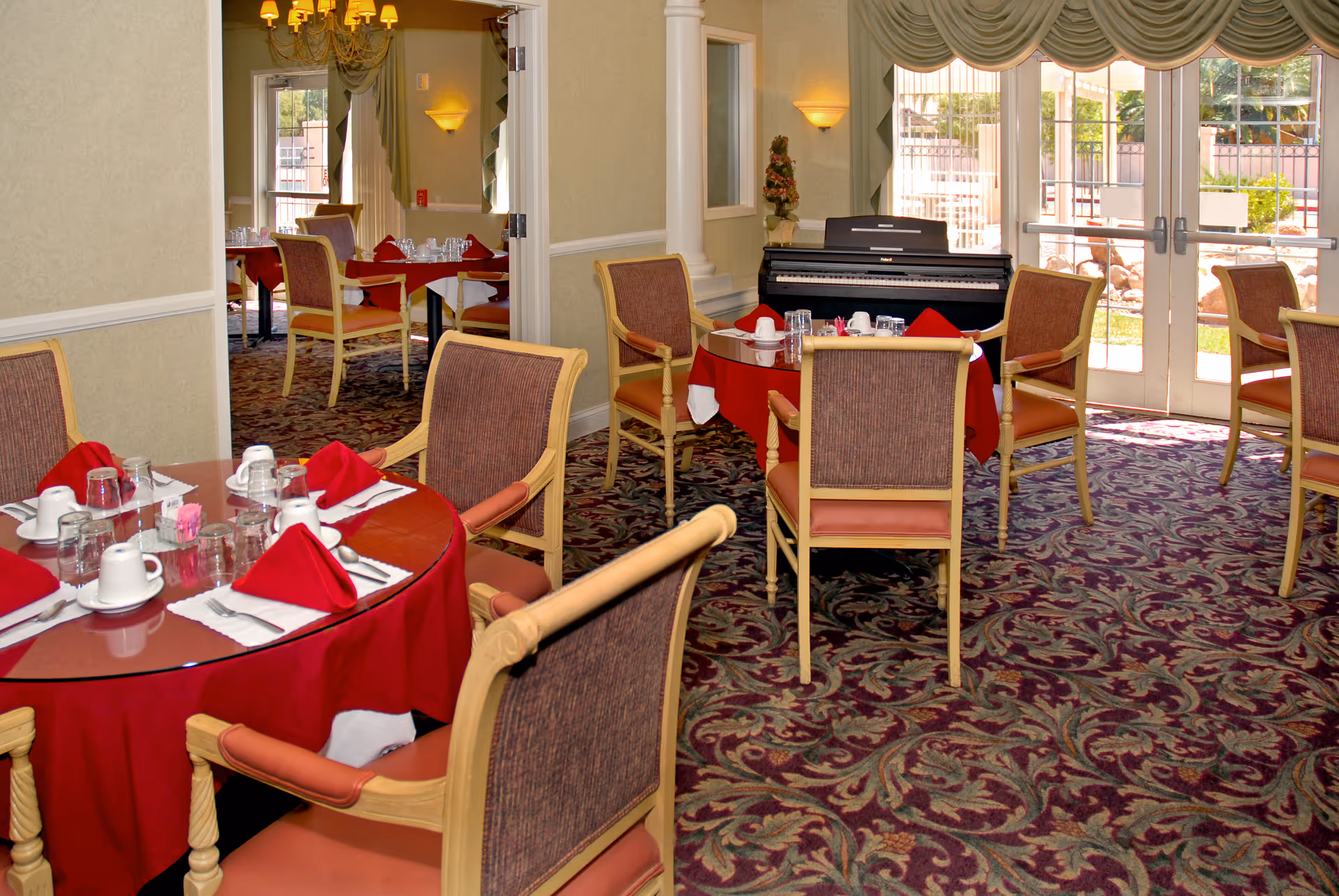 Dining room in a senior living facility with round tables covered in red tablecloths and set with white cups, glasses, and red napkins. The room has patterned carpet, wooden chairs with cushioned seats, a piano against the wall, and large windows with draped curtains letting in natural light.