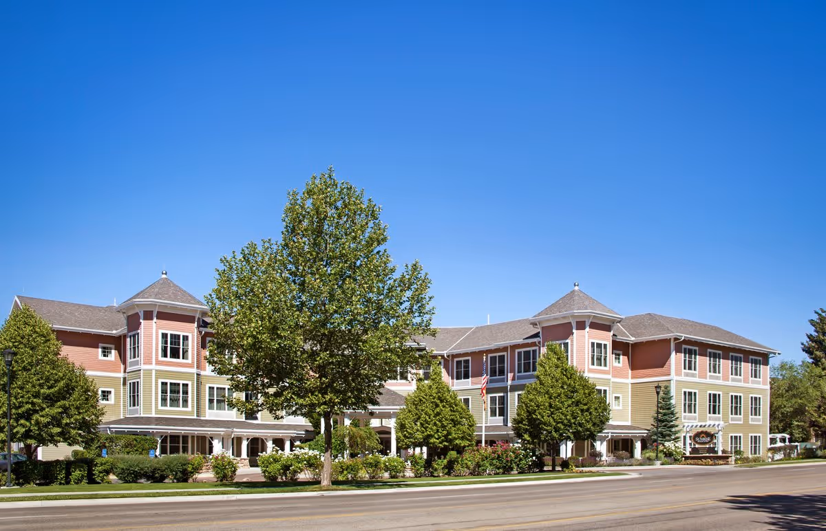Exterior view of a three-story senior living facility building with a mix of red and green siding, multiple windows, and a peaked roof. The building is surrounded by trees and landscaping, with a clear blue sky overhead.