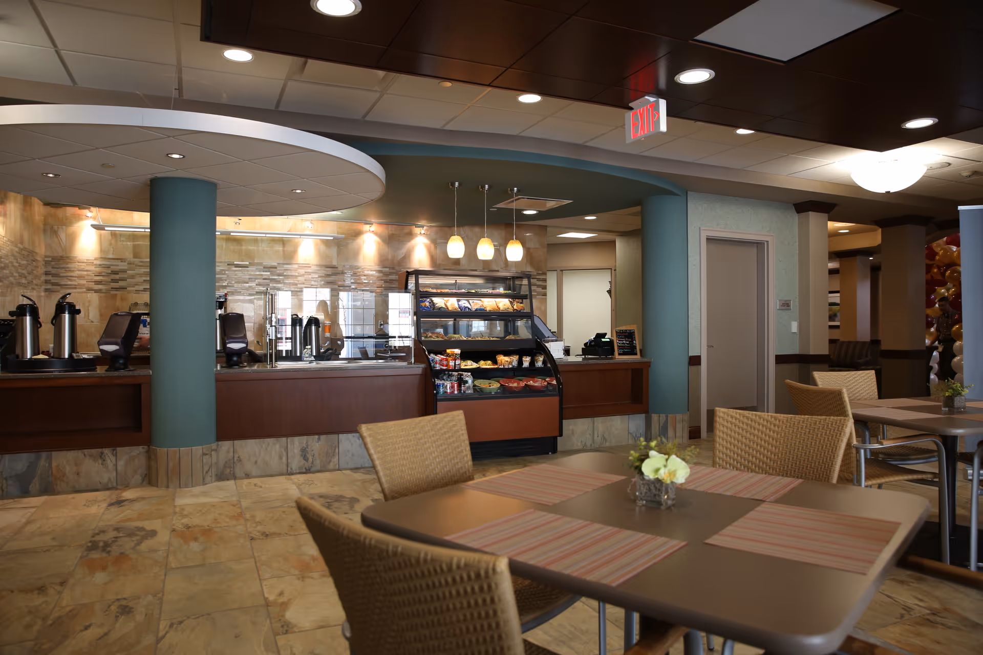 Interior view of a dining area in a senior living facility with tables and chairs. The foreground shows a table with four wicker chairs and a small flower arrangement. In the background, there is a counter with coffee dispensers and a display case with snacks and pastries, under hanging pendant lights. The floor is tiled and the ceiling has recessed lighting.