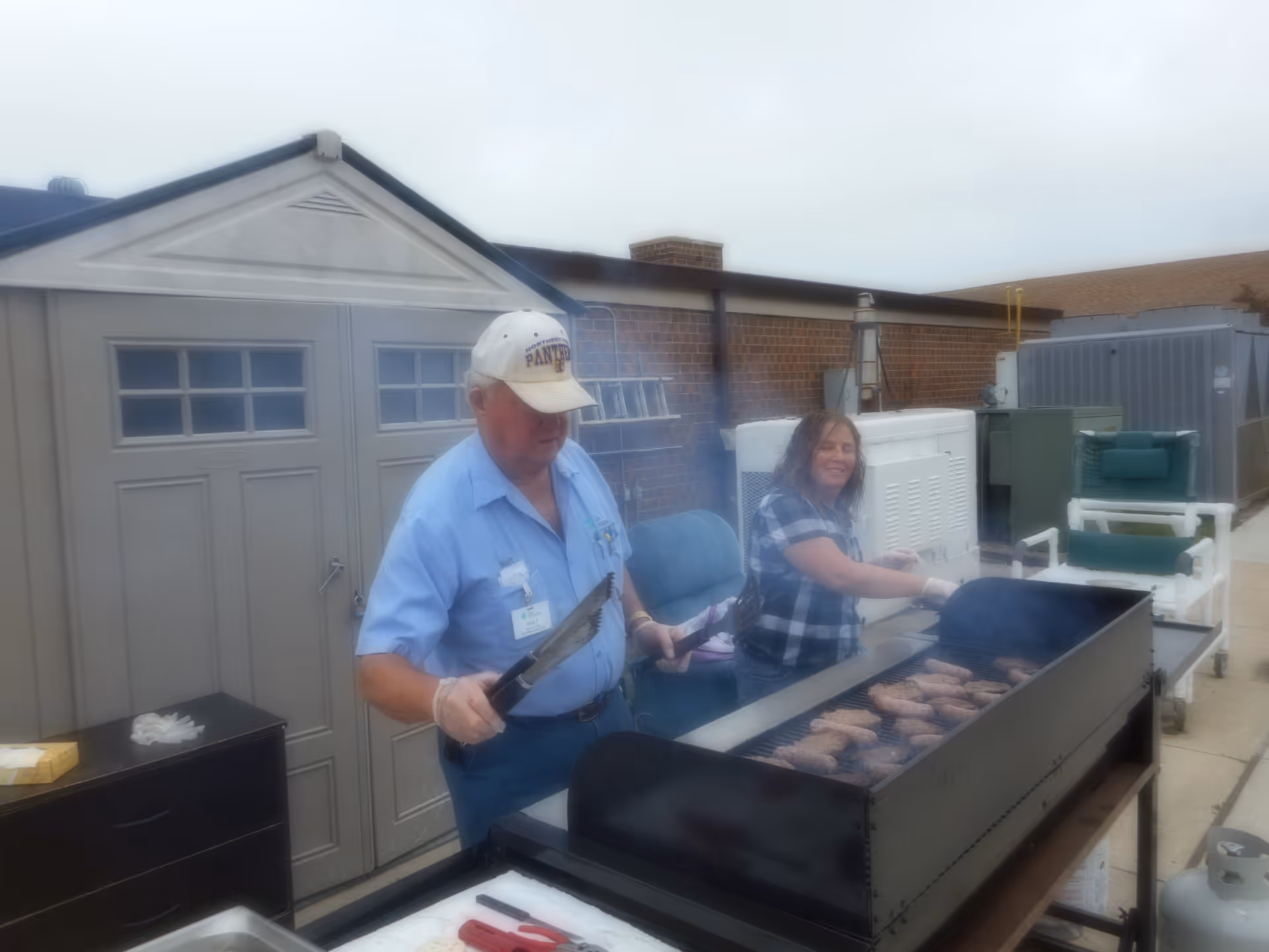 Two people cooking sausages on a large outdoor grill in a patio area with storage sheds and equipment in the background. One person is wearing a light blue shirt and a white cap, holding tongs, while the other person is smiling and tending the grill.