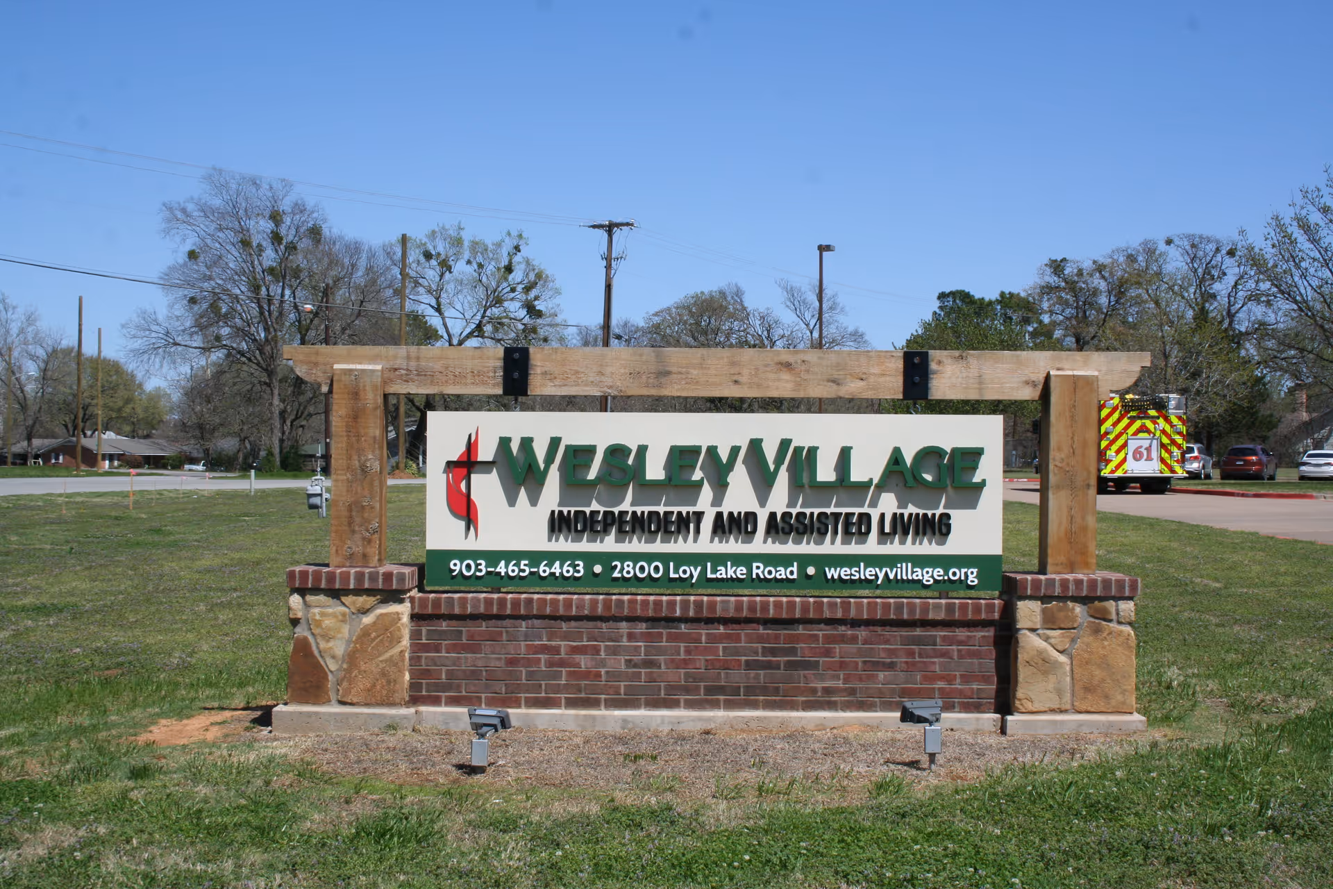 Outdoor sign for Wesley Village, an independent and assisted living facility, displayed on a brick and wood structure with a grassy area and trees in the background under a clear blue sky.