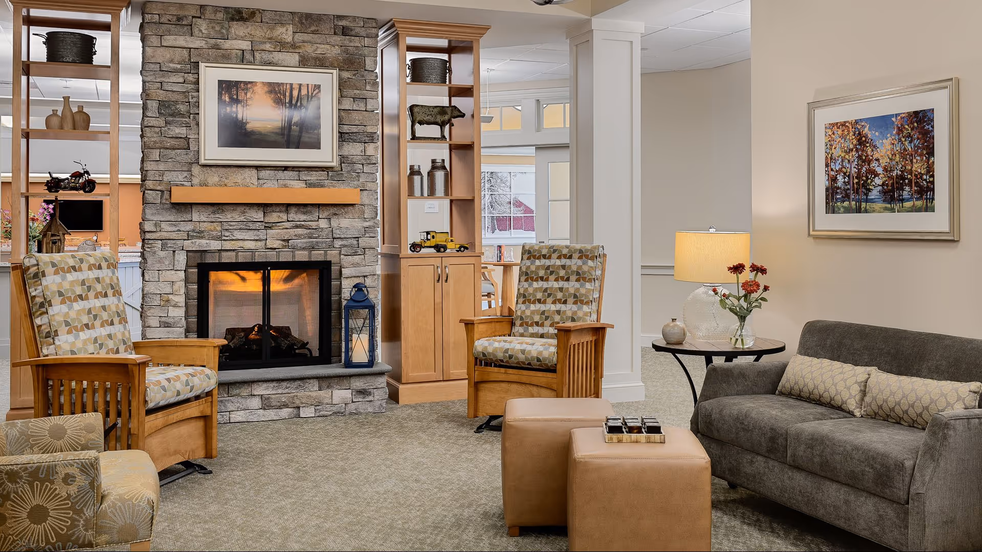 A cozy living room area with a stone fireplace in the center, flanked by two wooden armchairs with patterned cushions. To the right, there is a gray sofa with a decorative pillow, a round side table with a lamp and a vase of flowers. Two tan ottomans are placed in front of the sofa. The walls are adorned with framed artwork, and built-in wooden shelves with decorative items flank the fireplace.