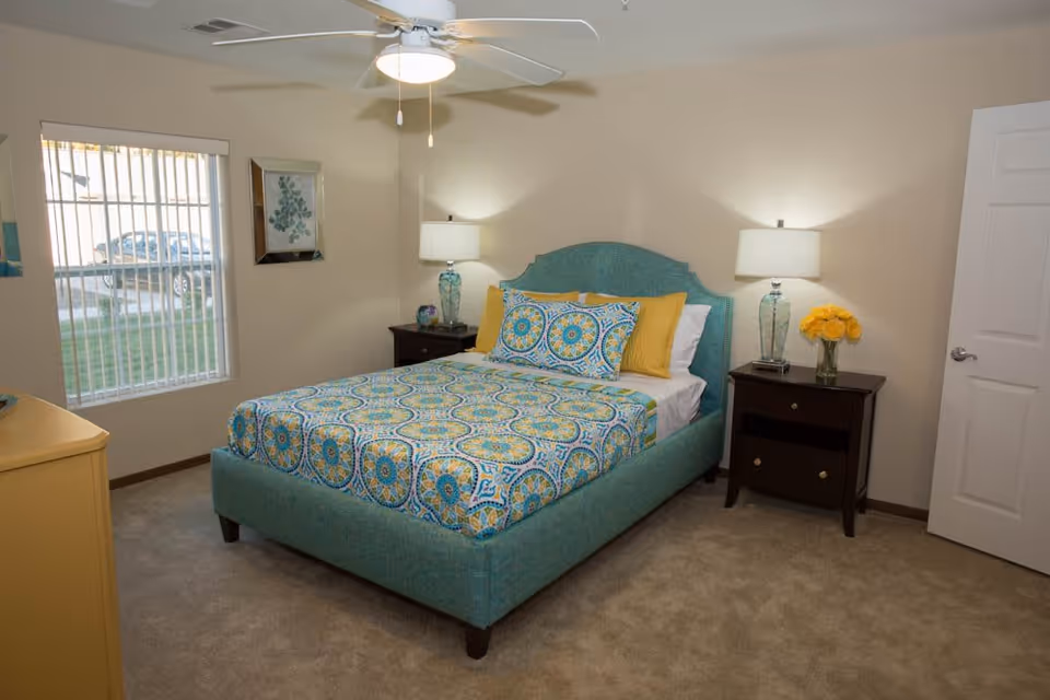 A neatly made bed with a turquoise upholstered headboard and a colorful patterned bedspread in shades of blue, yellow, and white. The room has beige walls, a ceiling fan with a light, two dark wooden nightstands with glass lamps and a vase of yellow flowers, a window with vertical blinds, and a closed white door.