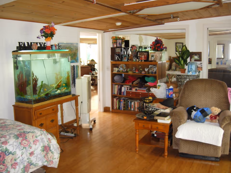 A cozy living room area with a brown armchair covered partially with a white cloth, a wooden coffee table with books and a fruit basket, a wooden bookshelf filled with books and decorative items, and a large fish tank on a wooden stand. The room has wooden flooring and a wooden ceiling, with a floral bedspread partially visible on the left side. There are flowers and framed photos on top of the bookshelf and fish tank, and a mirror on the wall reflecting part of the room.