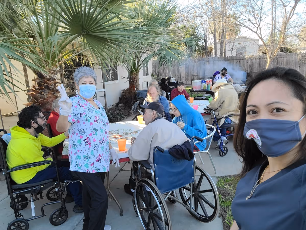 A group of elderly people and caregivers gathered outdoors at Rose Garden Assisted Living. Some elderly individuals are seated in wheelchairs around tables with colorful cups, while two caregivers wearing masks and gloves are standing and smiling at the camera. Palm trees and a wooden fence are visible in the background.