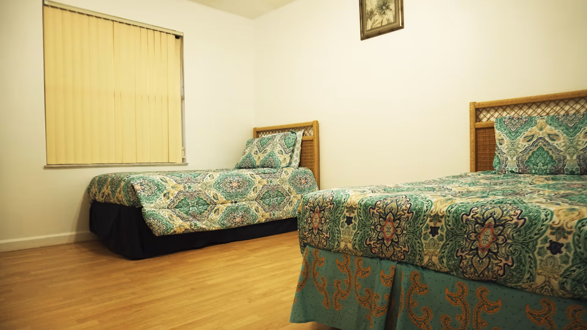 A simple bedroom with two twin beds featuring matching green and blue patterned bedspreads and pillows. The room has light-colored wooden flooring, a window with vertical blinds, and a framed picture hanging on the wall above one of the beds.