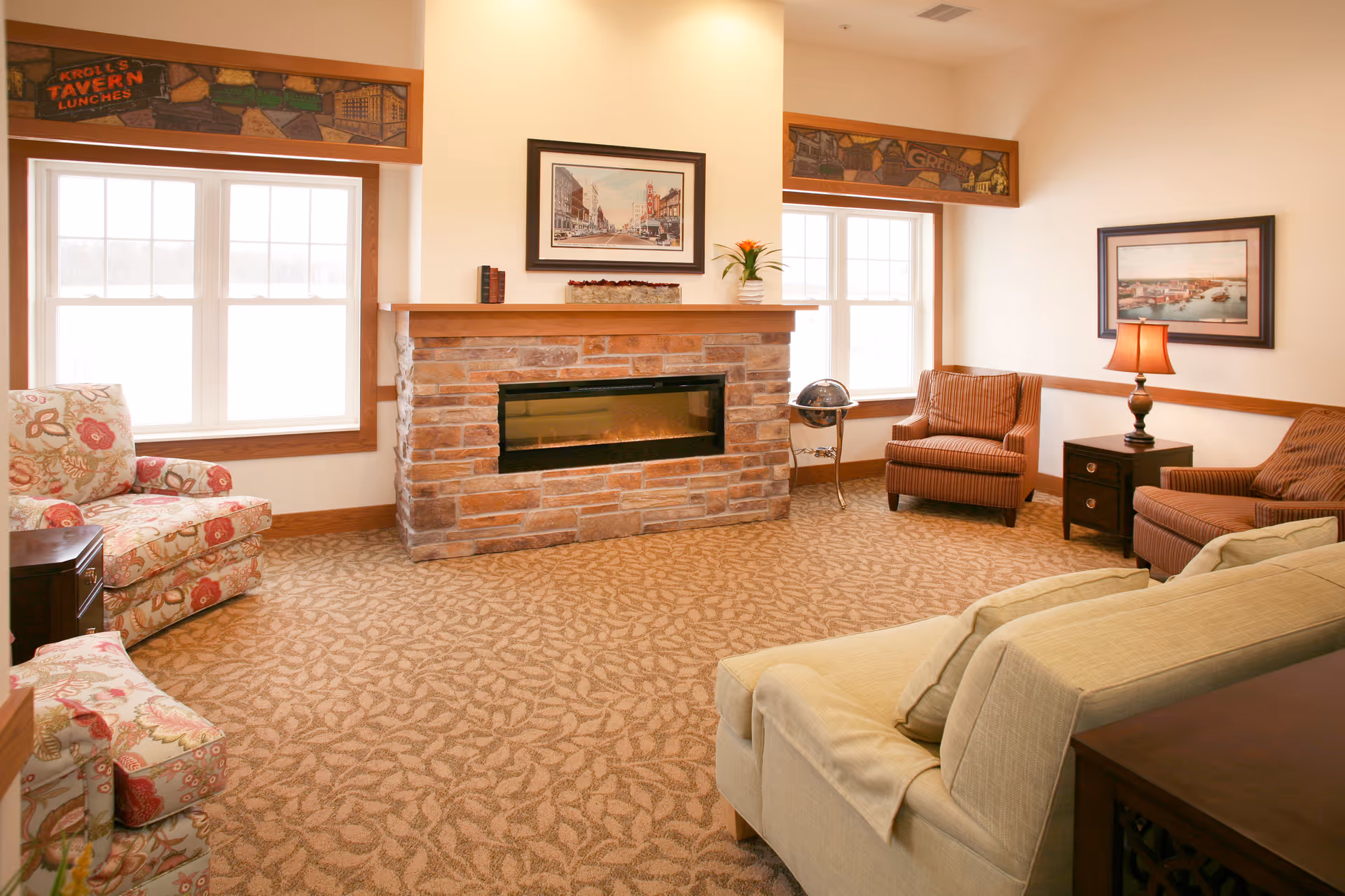 Bright common room with a stone fireplace, sofas and armchairs arranged on patterned carpet.