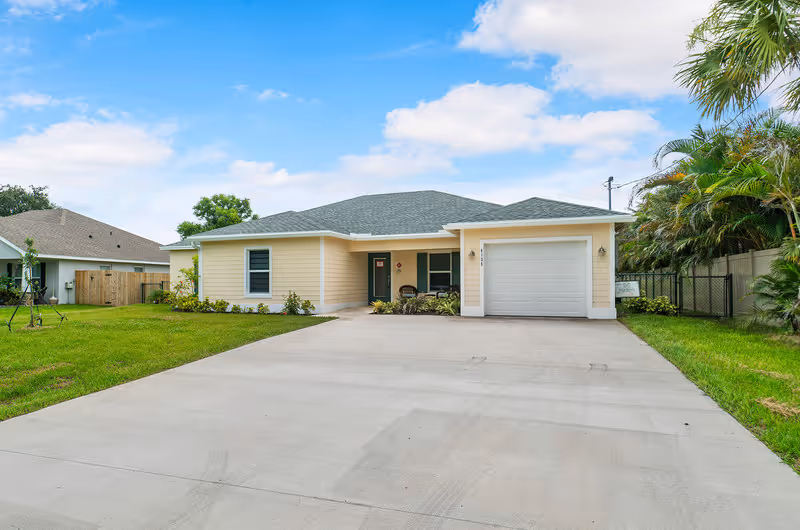 Single-story yellow house with a wide concrete driveway, attached garage, and small front porch surrounded by lawn and palm trees.