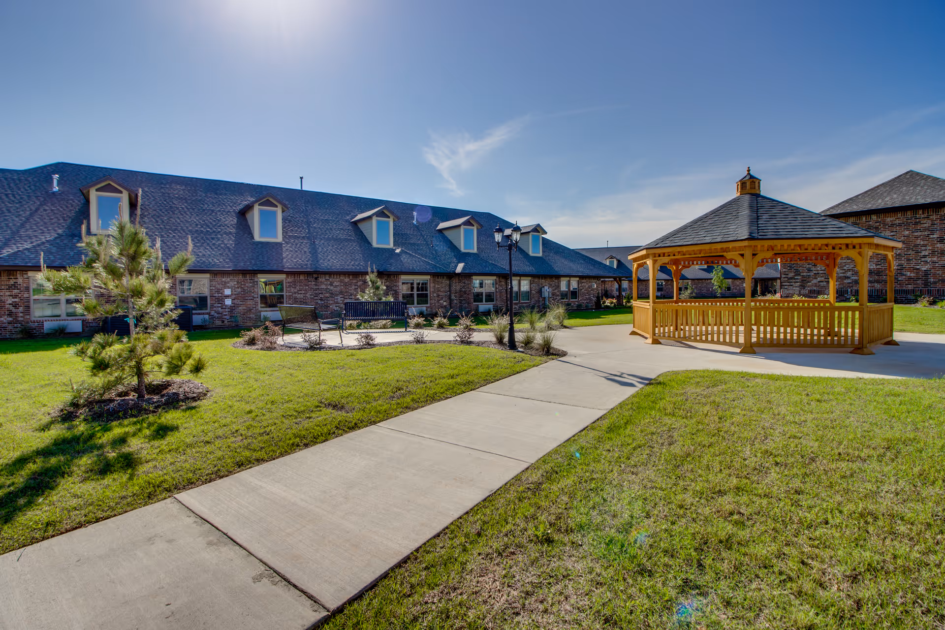 Outdoor courtyard area of Mayberry Gardens Assisted Living and Memory Care Homes featuring a paved walkway, green grass, small trees, benches, a street lamp, and a wooden gazebo under a clear blue sky.