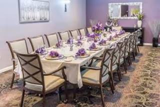 A long dining table set for a formal meal with white tablecloth, purple napkins, and glassware in a room with lavender walls, a large mirror, and floral carpet.