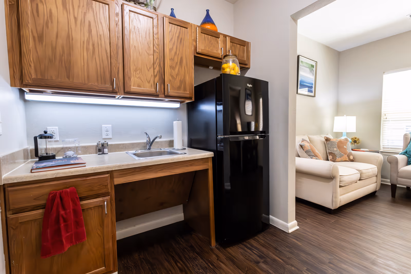 A small kitchen area with wooden cabinets, a countertop with a sink, a black refrigerator, and a red towel hanging from a cabinet handle. Adjacent to the kitchen is a living room with a beige loveseat, a lamp on a side table, and a window with blinds.