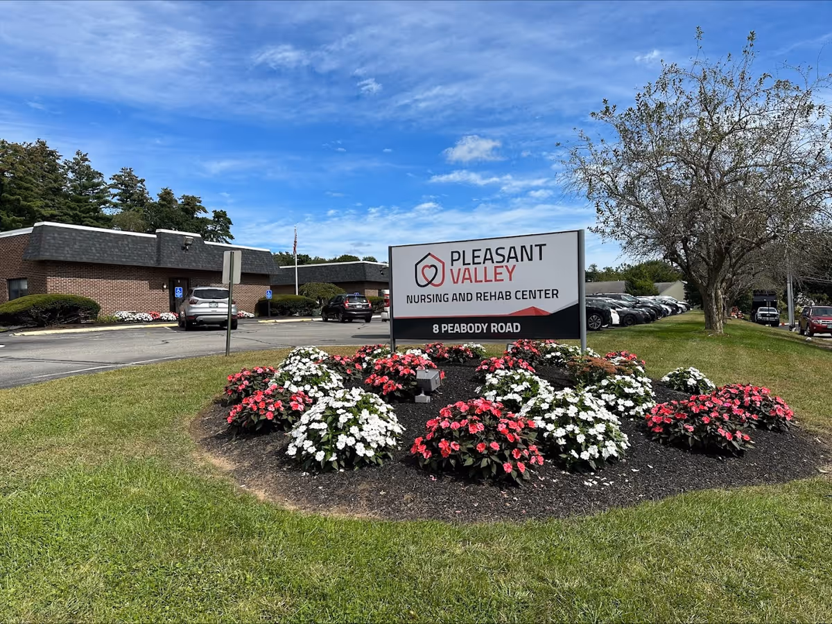 Outdoor view of Pleasant Valley Nursing and Rehab Center with a large sign displaying the facility name and address, surrounded by flower beds with pink and white flowers. The building and parking lot with several cars are visible in the background under a partly cloudy blue sky.