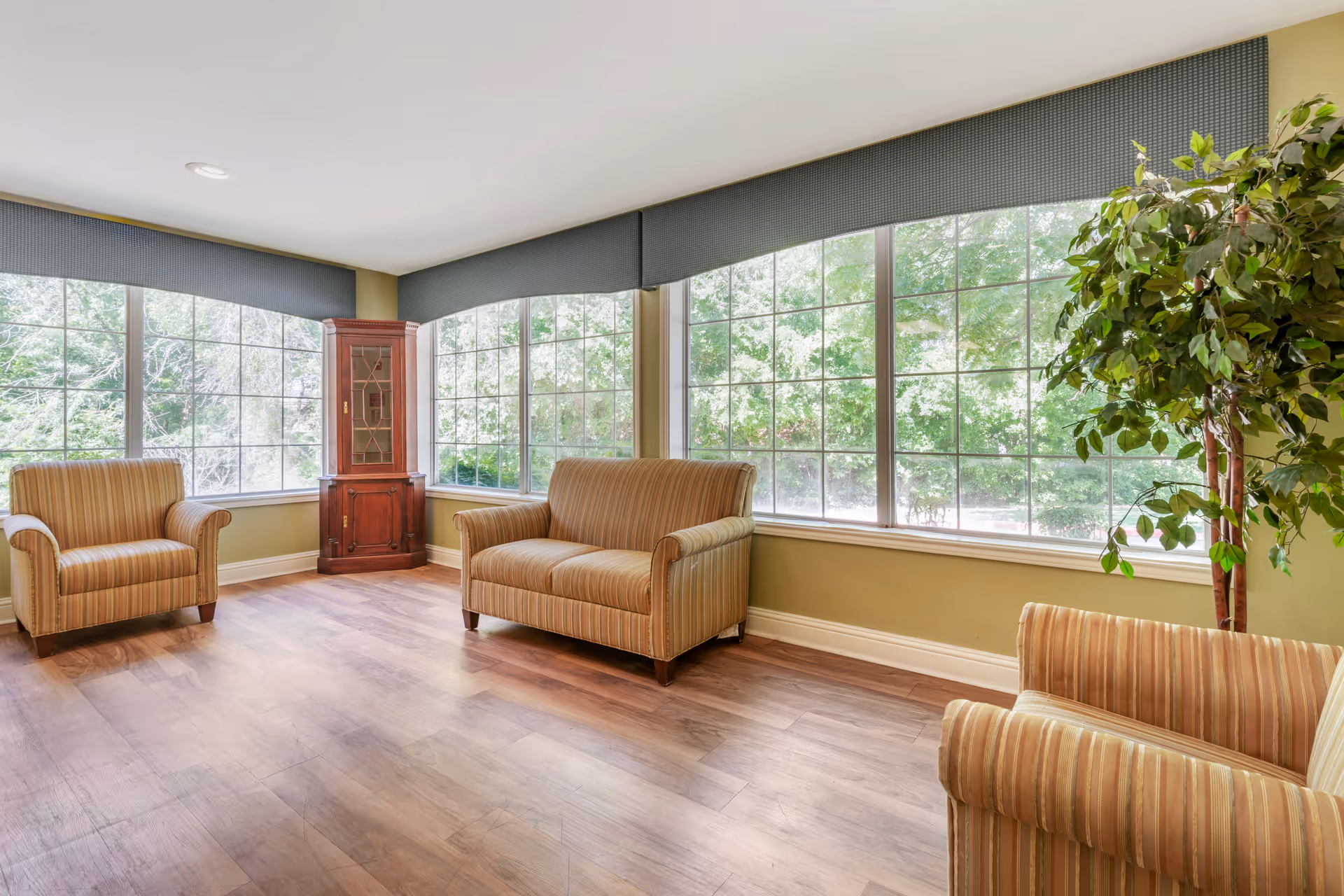 A bright common room with striped armchairs and a loveseat facing large grid windows, a wooden corner cabinet, and a potted plant.