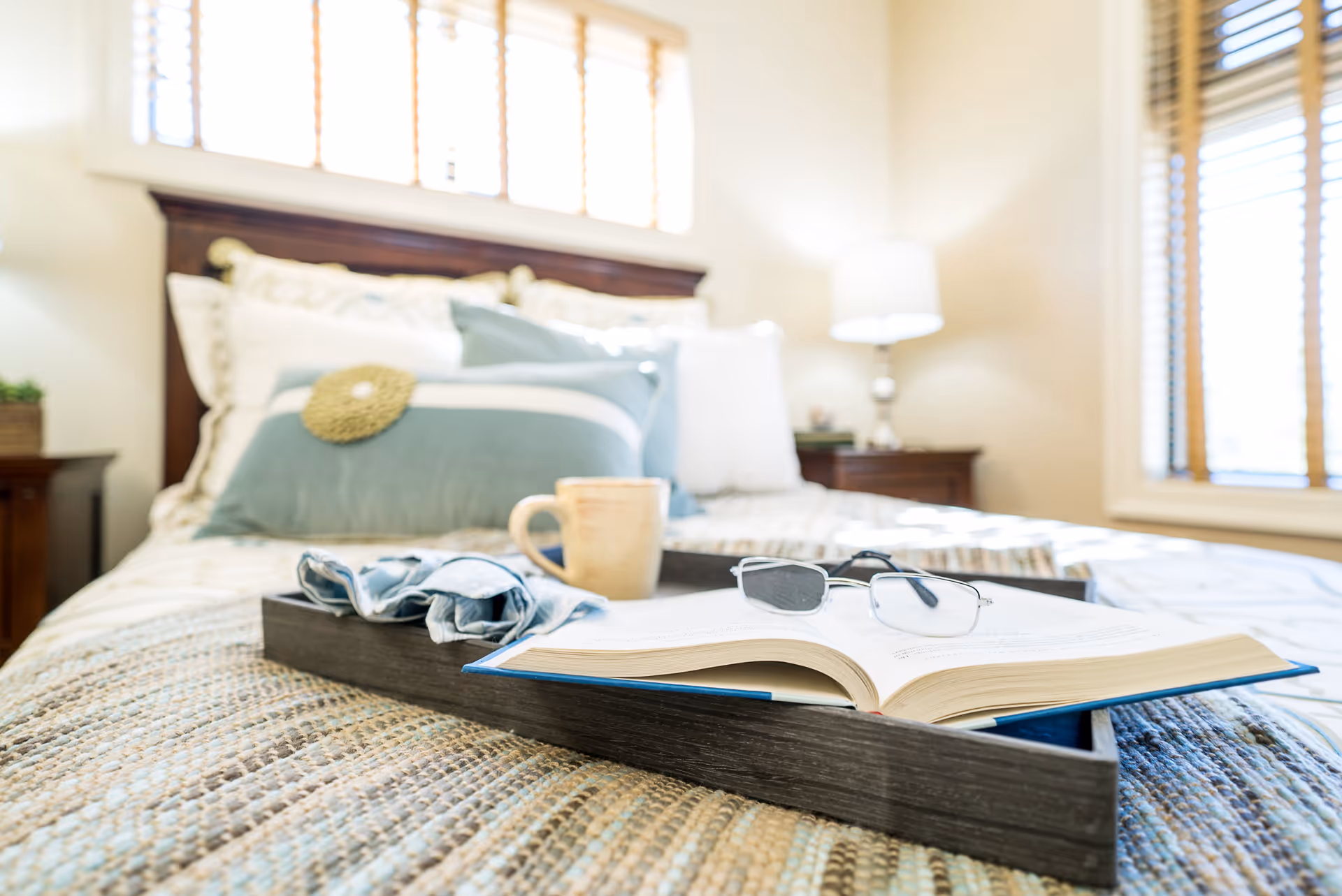 A cozy bedroom with a neatly made bed featuring multiple pillows in white and blue tones. On the bed is a dark wooden tray holding an open book with reading glasses resting on it, a ceramic mug, and a folded cloth. The room is softly lit by natural light coming through windows with wooden blinds and a bedside lamp on a wooden nightstand.