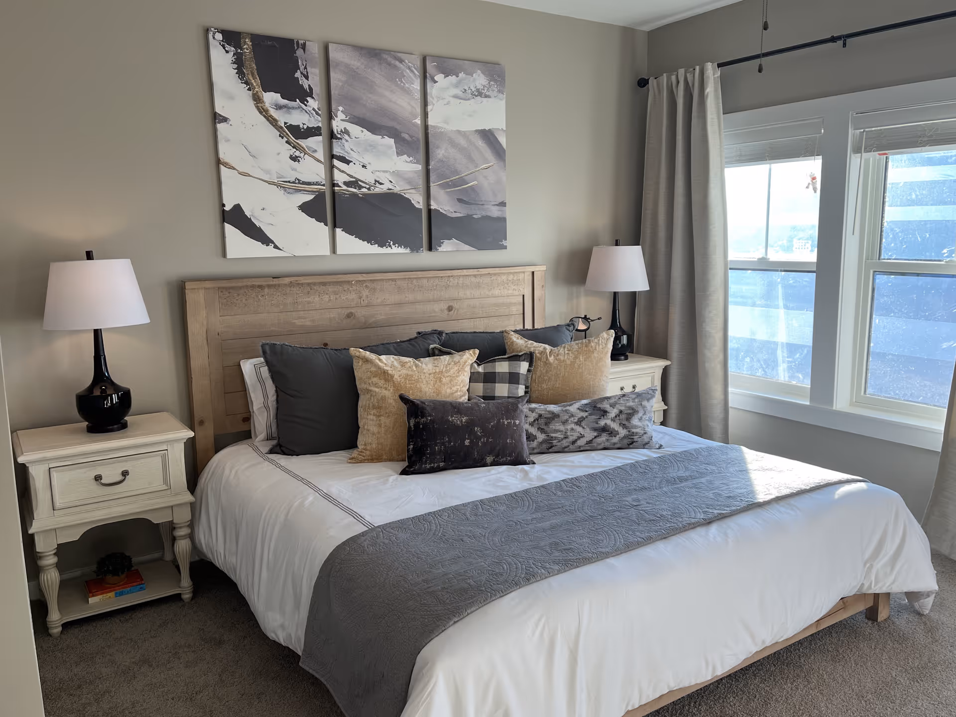 A cozy bedroom with a wooden headboard bed adorned with multiple pillows in shades of gray, beige, and black. There are two matching bedside tables, each with a black lamp with a white lampshade. Above the bed, there is a three-panel abstract painting in black, white, and gray tones. The room has beige walls, carpeted floor, and a window with light gray curtains letting in natural light.