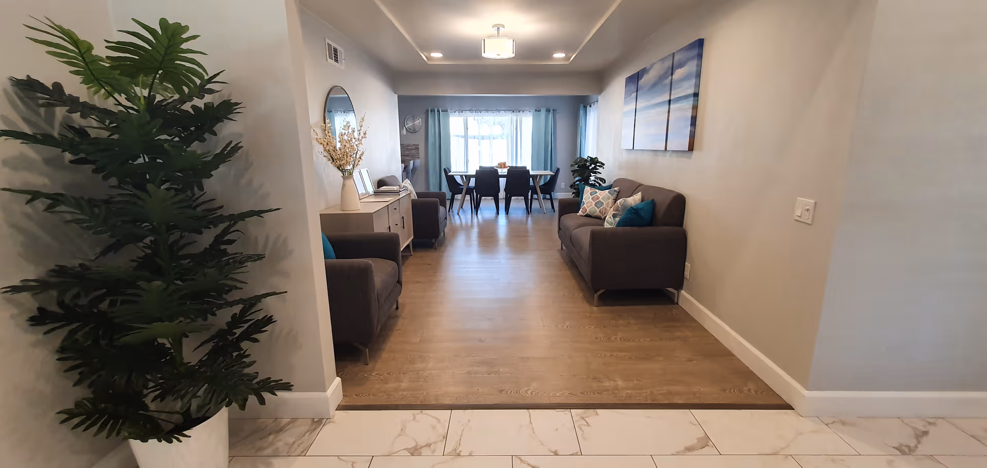 Interior view of a senior living facility showing a hallway leading to a living room and dining area. The foreground has a large potted plant on the left and tiled flooring transitioning to wooden flooring. The living room features two gray sofas with colorful cushions, a sideboard with decorative items, and a round mirror on the wall. The dining area at the far end has a table with six chairs and a large window with curtains letting in natural light.