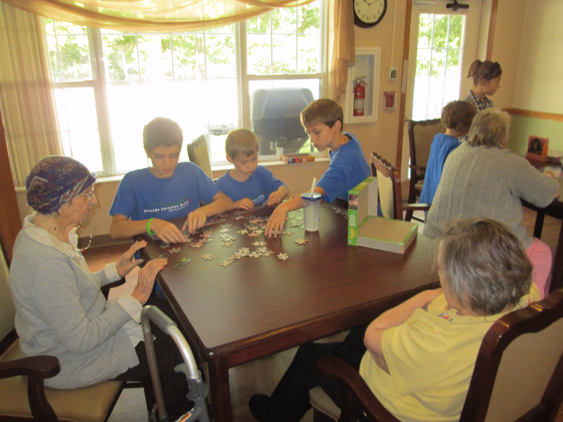 A group of elderly people and children sitting around a wooden table working on a jigsaw puzzle together in a well-lit room with large windows. The elderly individuals are seated in chairs, and one has a walker nearby. The children are wearing matching blue t-shirts.