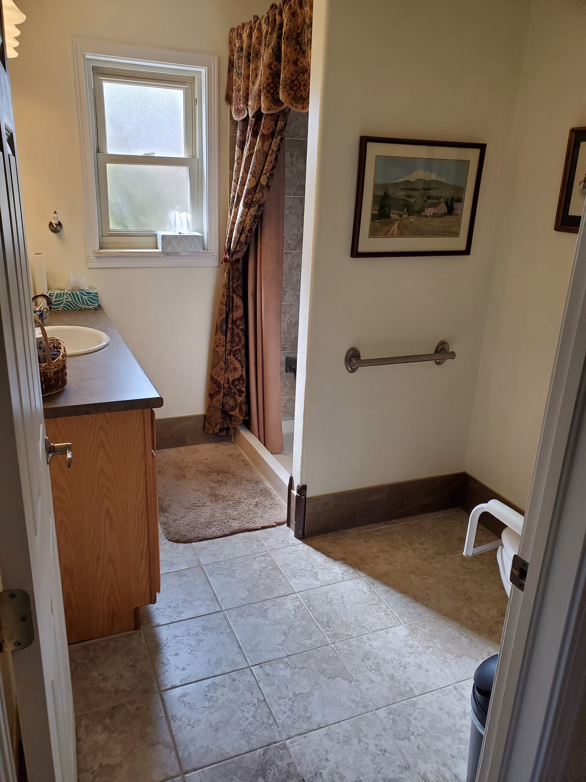 A bathroom with a tiled floor, a wooden vanity with a sink on the left, a window above the vanity, a shower area with a floral curtain, a brown bath mat, a grab bar on the wall, and framed artwork hanging above the grab bar.
