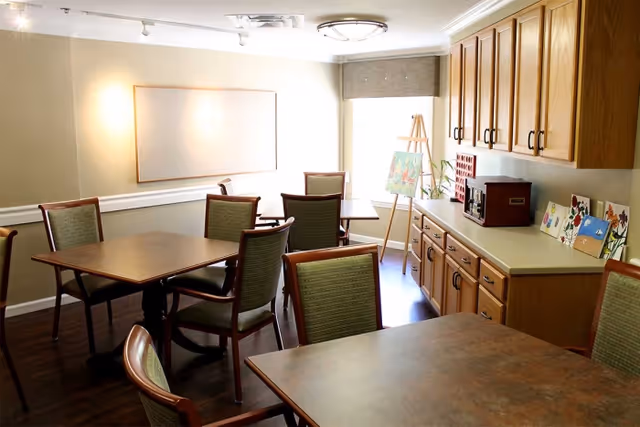 A bright multipurpose dining/activity room with several tables and green-upholstered chairs, wooden cabinets, and an easel displaying paintings.