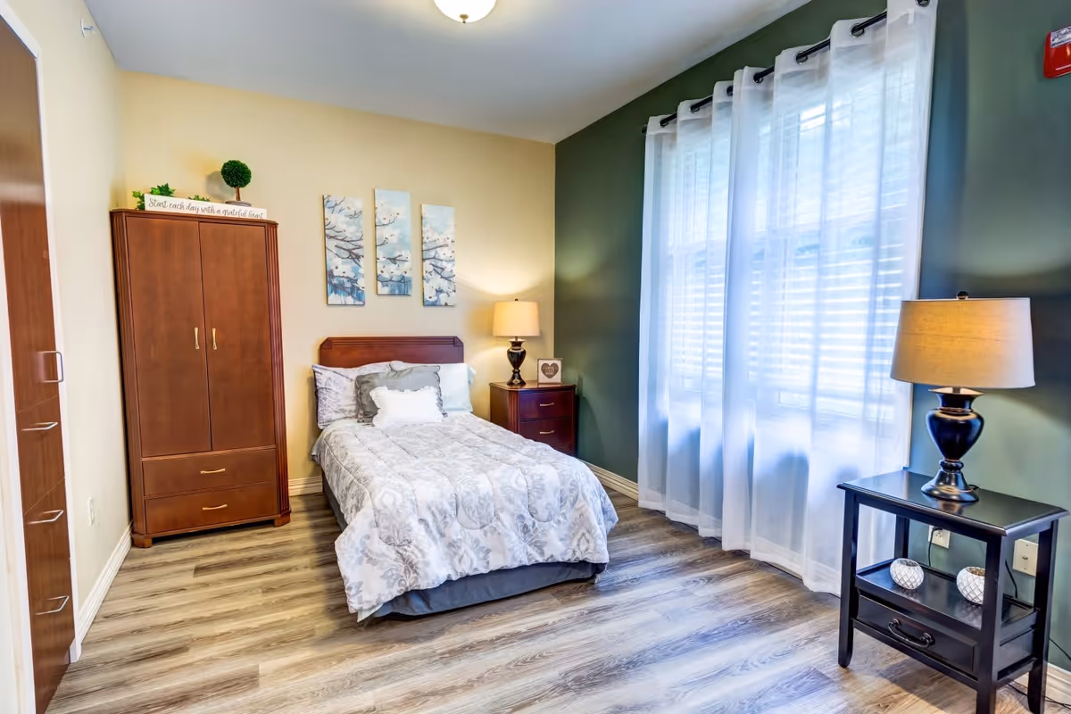 A cozy bedroom with a single bed covered in a patterned comforter and pillows. There is a wooden wardrobe on the left side, a nightstand with a lamp and decorative items next to the bed, and a small black table with a lamp and decorative objects near a large window with sheer white curtains. The walls are painted in two tones, beige and green, and the floor has wood-like vinyl planks.