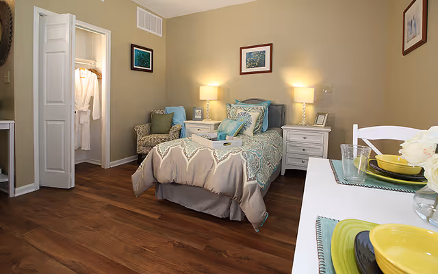 Furnished bedroom with a single bed, two nightstands with lamps, an armchair, open closet, and a small dining surface.