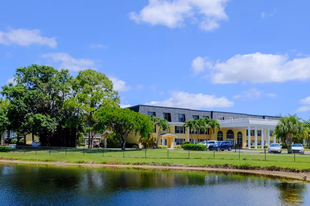 Exterior view of Volante Senior Living of Delray Beach, a yellow building with white columns and a black roof, surrounded by trees and a small pond in the foreground under a blue sky with scattered clouds.