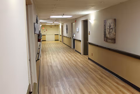 A long, well-lit hallway in a nursing and rehabilitation facility with light wood flooring, beige walls with a darker beige lower half, handrails on both sides, wall-mounted lights, and decorative artwork. Several doors line the hallway, and an exit sign is visible at the far end.