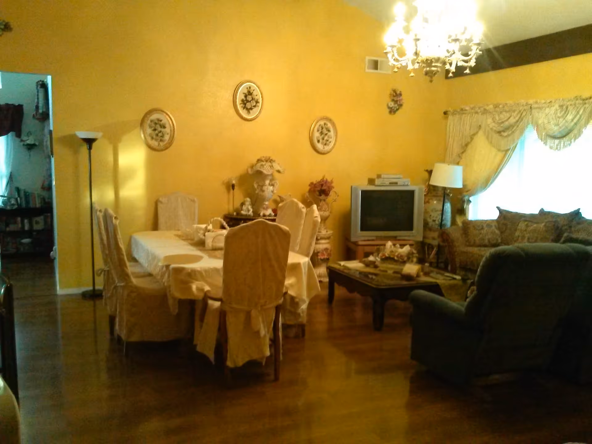 A cozy living and dining area with yellow walls. The dining area has a table covered with a white tablecloth and six chairs with white covers. The living area features a sofa, an armchair, a coffee table with decorative items, a TV on a stand, and a floor lamp. The room is decorated with floral wall hangings and a chandelier, with a window covered by ornate curtains.