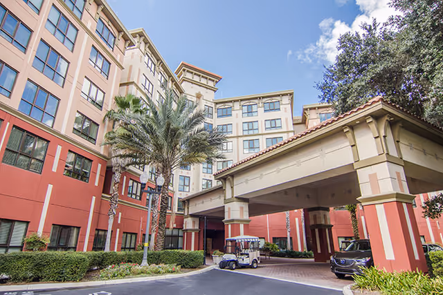 Exterior view of a multi-story senior living facility with a covered entrance, palm trees, and parked vehicles including a golf cart and a car under the canopy. The building has large windows and a mix of beige and red exterior walls under a partly cloudy sky.