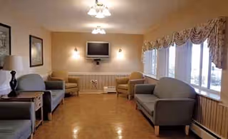 Well-lit senior living lounge with blue sofas, chairs, and a wall-mounted TV.