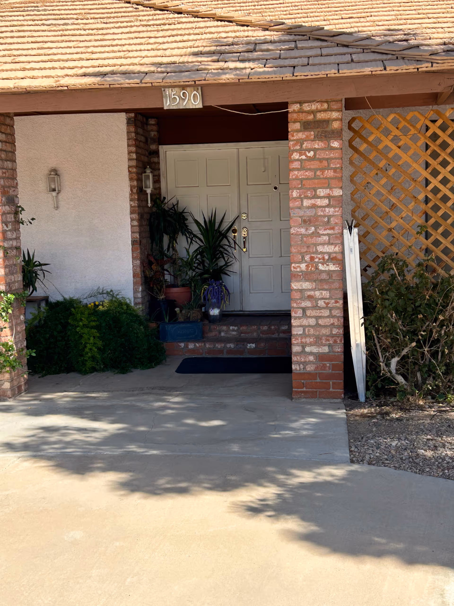 Front entrance of a building with a covered porch supported by brick columns. The door is white with two panels and has gold-colored handles. There are several potted plants on the porch and a lattice panel on the right side. The address number 1590 is displayed above the door.