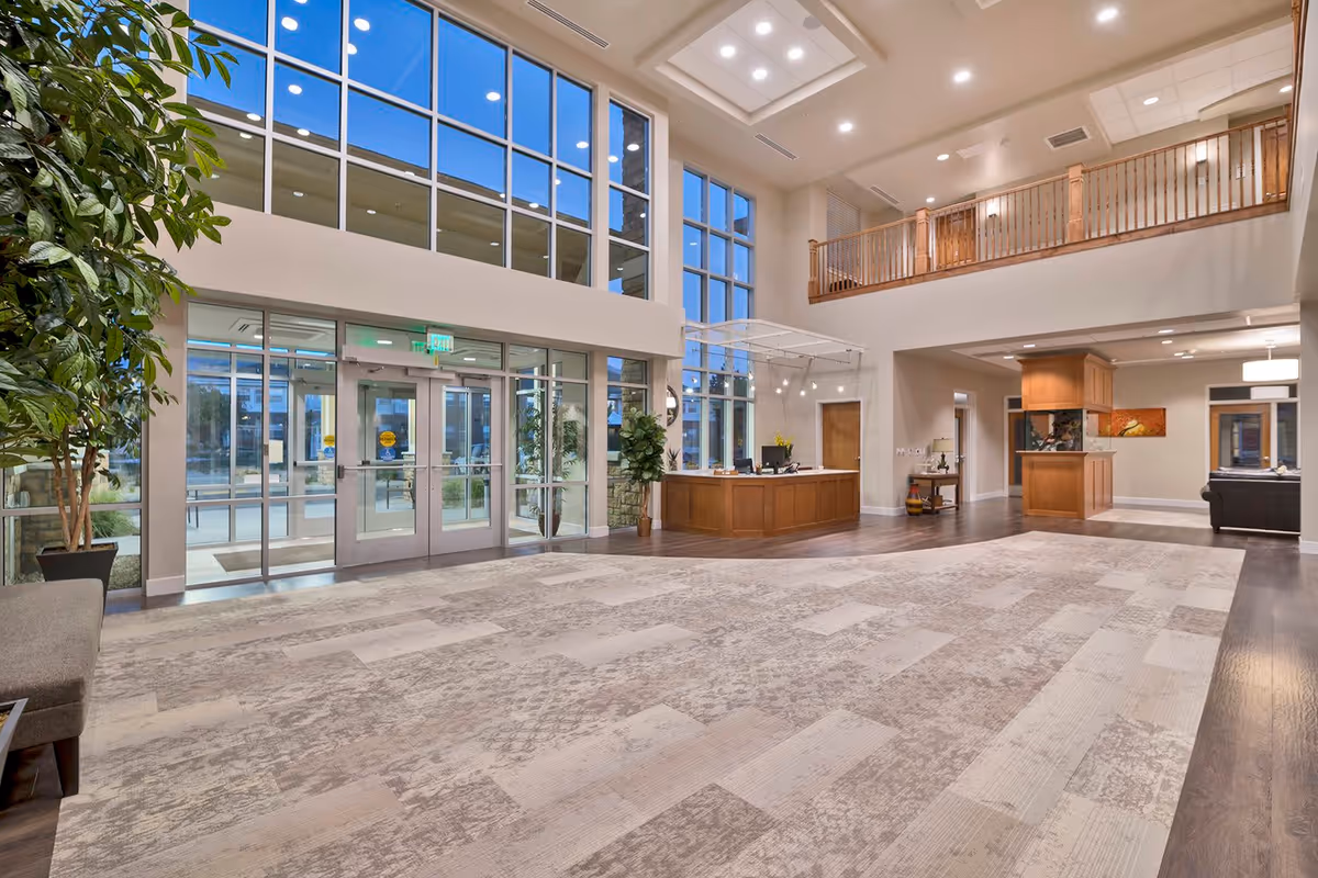 Spacious and well-lit senior living facility lobby with large floor-to-ceiling windows, a reception desk, indoor plants, and a second-floor balcony overlooking the area.