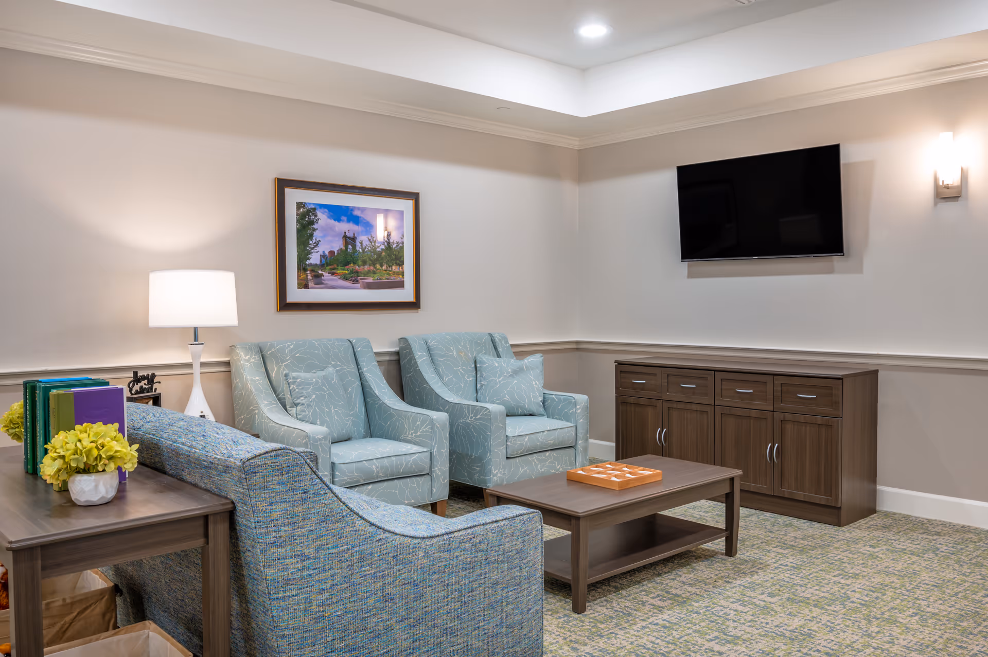 A cozy living room area with two light blue patterned armchairs, a matching sofa, a wooden coffee table with a game on it, a wooden sideboard, a wall-mounted flat screen TV, a table lamp, and a framed picture on the wall.