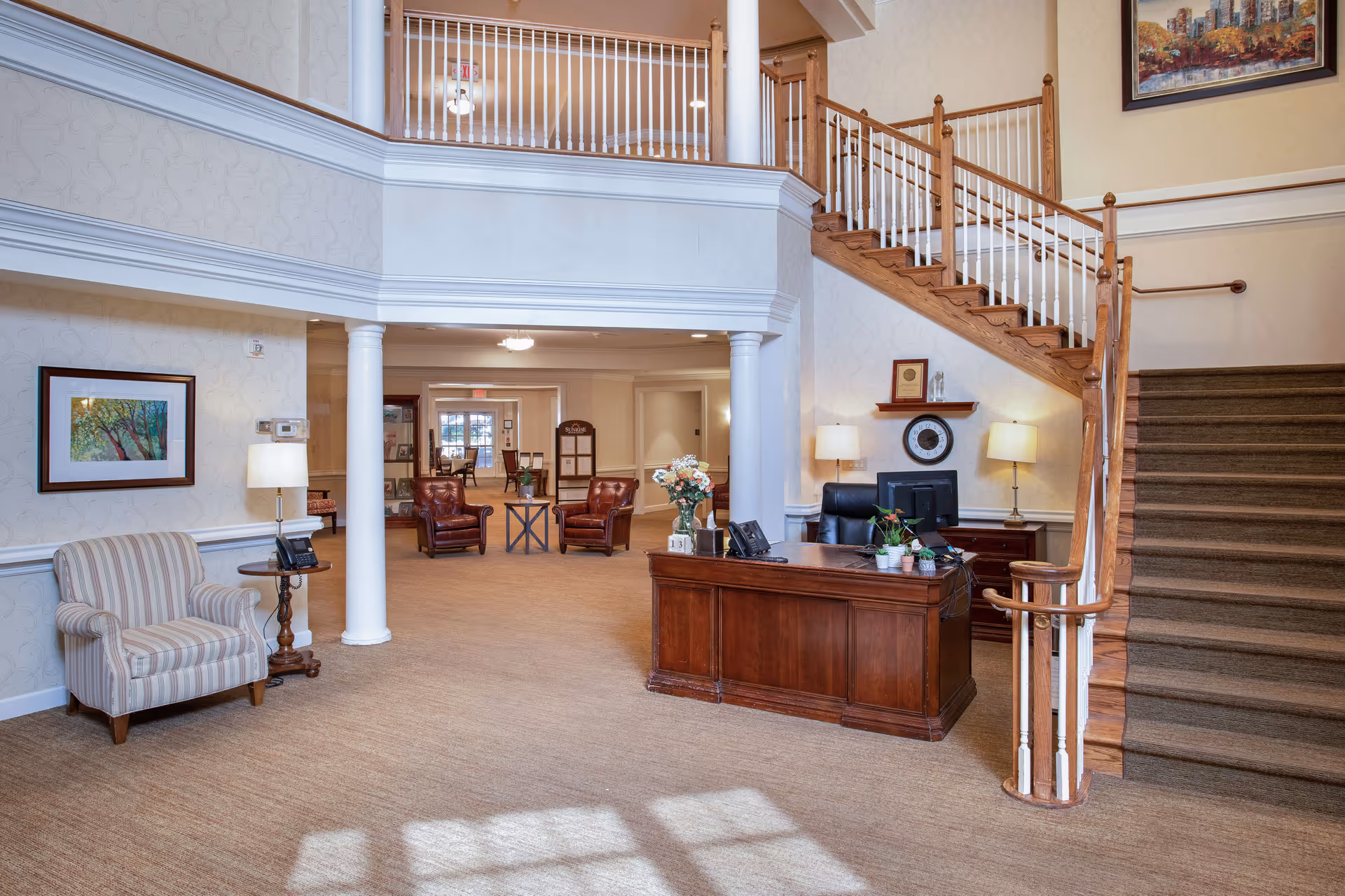 A spacious and well-lit reception area in a senior living facility featuring a wooden reception desk with a computer and plants, a staircase with wooden banisters leading to an upper floor, two white columns, a striped armchair with a side table and lamp, and a seating area with leather chairs and a small table in the background.