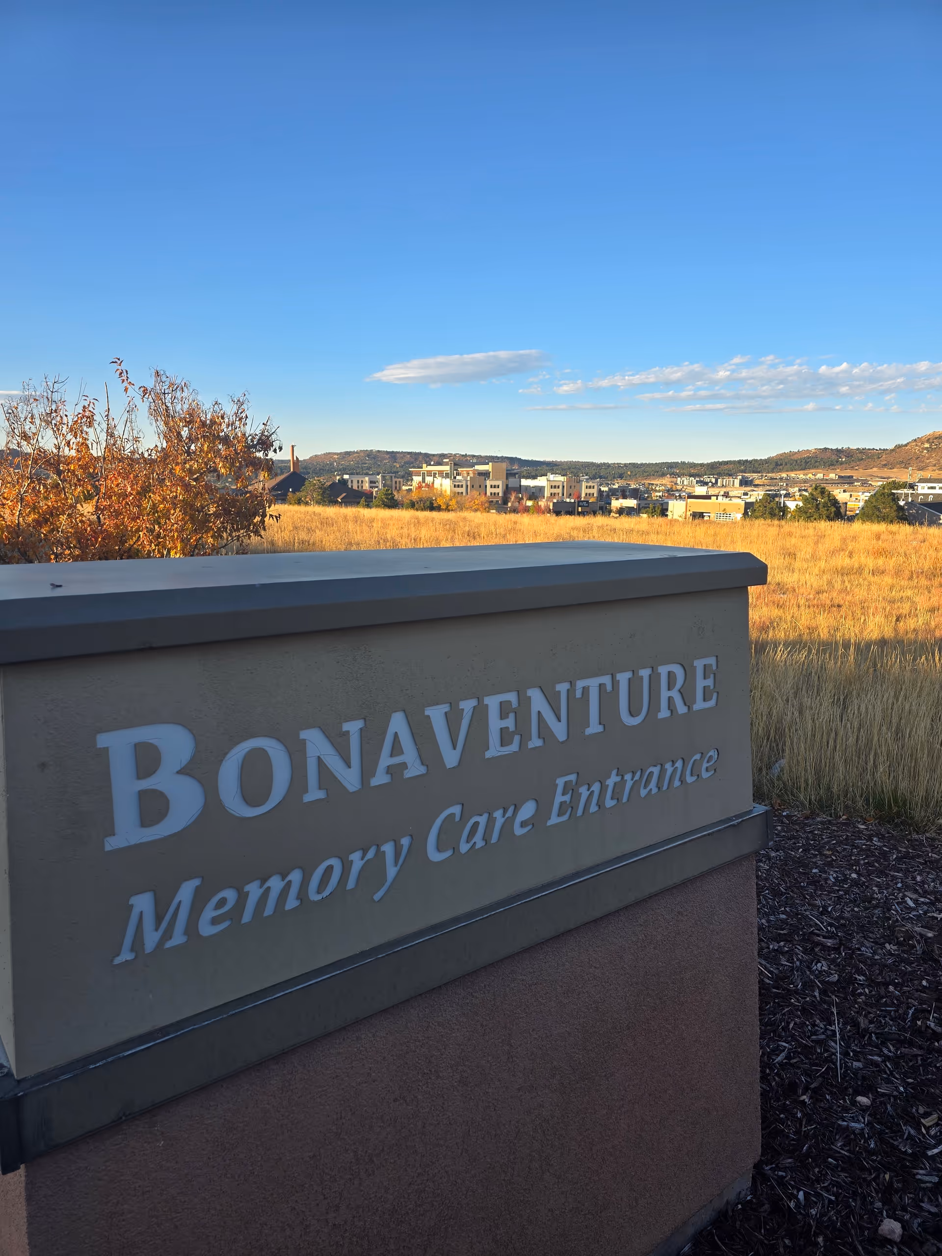 Outdoor sign for Bonaventure Memory Care Entrance with a field and buildings in the background under a clear blue sky.