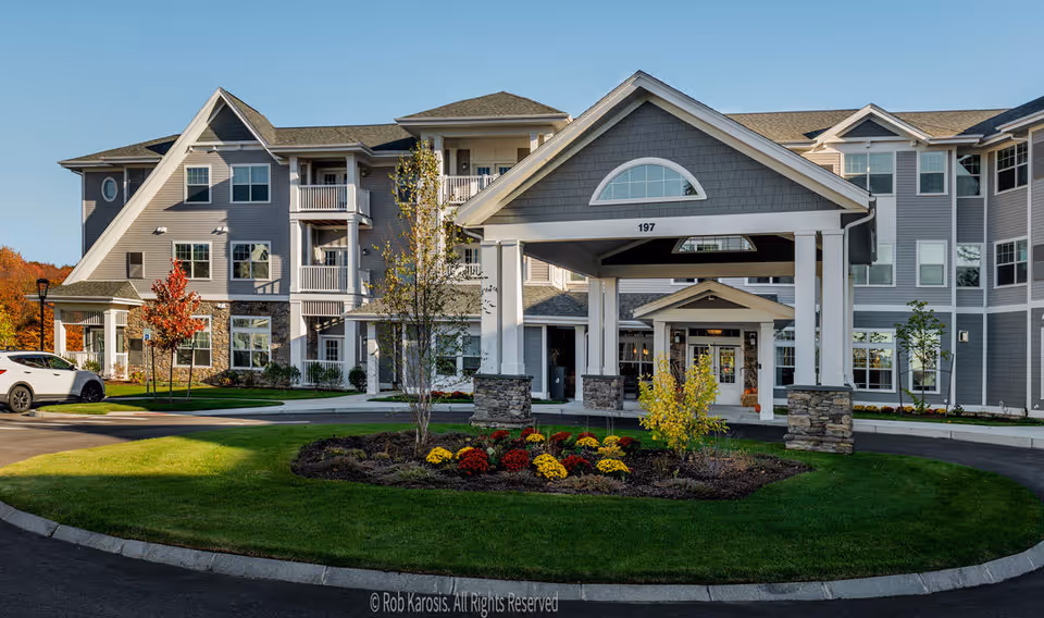 Front exterior view of a large, multi-story retirement community building with gray siding and white trim. The entrance features a covered porte-cochère with stone pillars and a landscaped roundabout with colorful flowers and small trees. A white car is parked on the left side near the building.