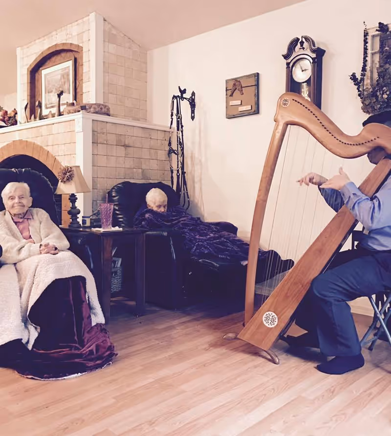 Two elderly women sitting in armchairs in a cozy living room with blankets, while a person plays a large wooden harp. The room features a tiled fireplace, a grandfather clock, and wooden flooring.