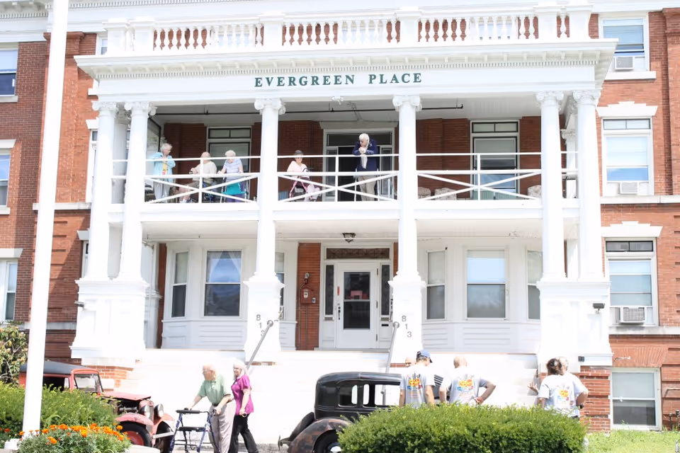 Front exterior view of Evergreen Place, a large brick building with white columns and a balcony. Several elderly people are standing and sitting on the balcony, and a few people are gathered near vintage cars and bushes in front of the building.
