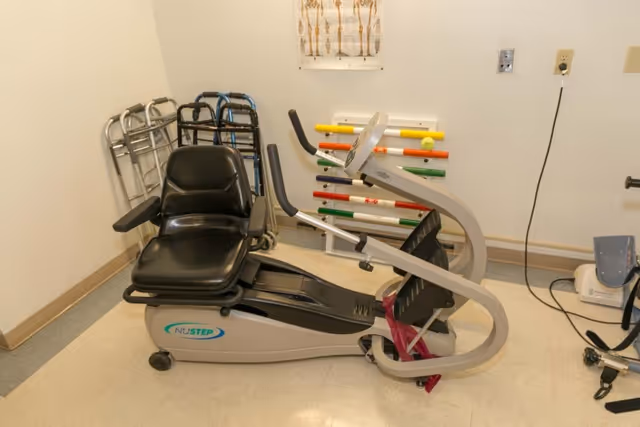 A small room with a NuStep exercise machine designed for seated use, several walkers lined up against the wall, a colorful pegboard mounted on the wall, and a medical device on the floor. The room has plain white walls and a tiled floor.