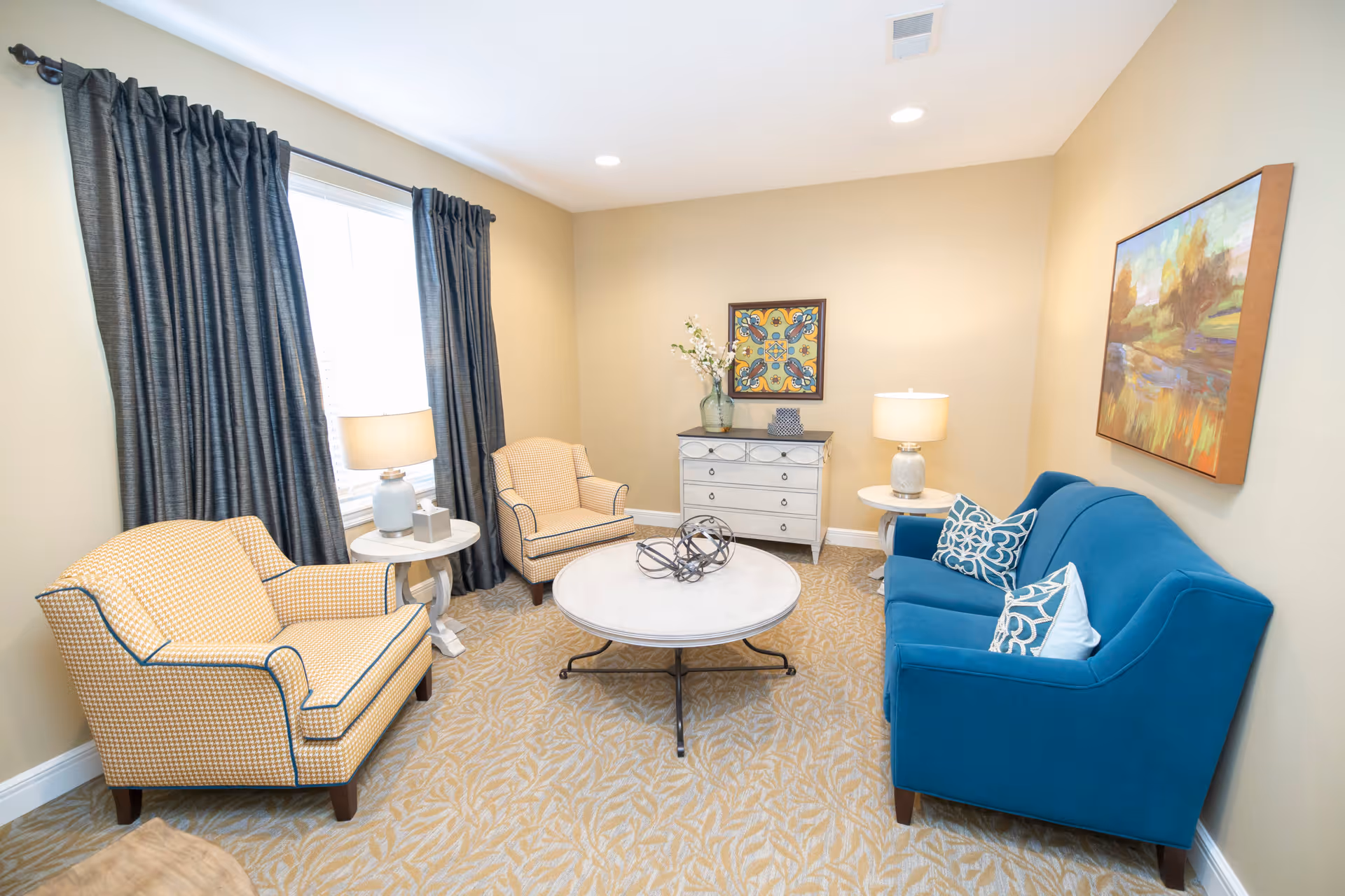 A cozy living room with a blue sofa, two beige armchairs, a round white coffee table with decorative items, two side tables with lamps, a small white dresser with a vase and decor, and two framed paintings on beige walls. A window with dark curtains lets in natural light.