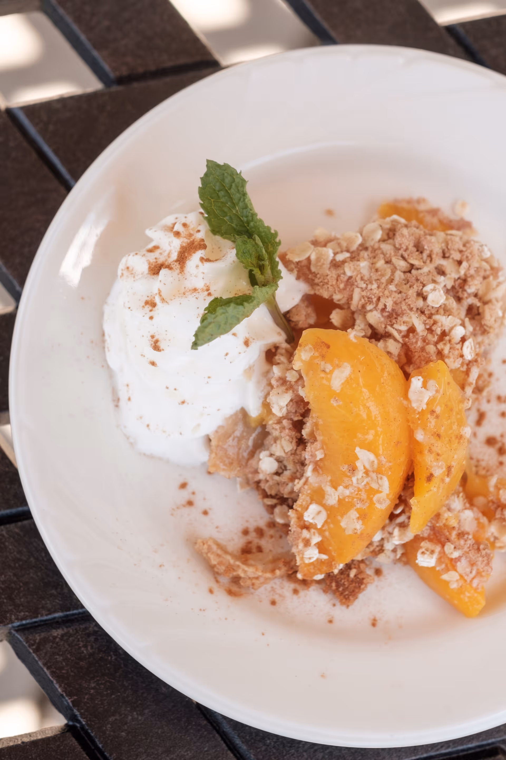 A white plate with a serving of peach crumble topped with oats and cinnamon, accompanied by a dollop of whipped cream garnished with a fresh mint leaf, placed on a dark slatted surface.