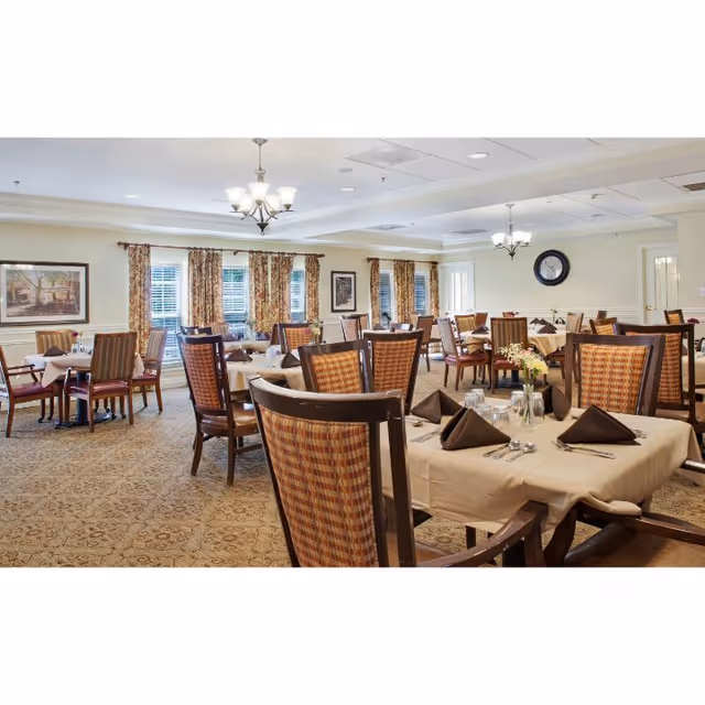 A spacious dining room with multiple tables covered in beige tablecloths, each set with brown folded napkins, silverware, and glassware. The room has patterned carpet, large windows with floral curtains, framed artwork on the walls, and two chandeliers hanging from the ceiling.