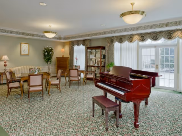 A spacious, well-lit living room with a polished grand piano and matching bench in the foreground. The room features patterned carpet, several wooden chairs and tables, a sofa, a tall cabinet with glass doors, a potted plant, and large windows with sheer curtains and decorative valances allowing natural light to fill the space.