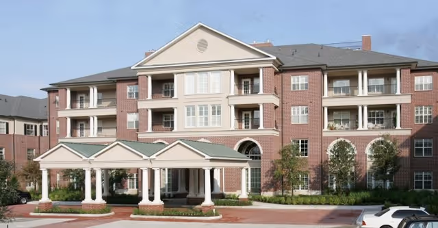 Front exterior view of a large, multi-story brick building with white columns and balconies, featuring a covered entrance with a green roof and landscaped surroundings under a clear sky.