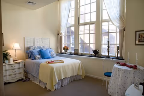 Bright bedroom with a made bed beside a large multi-pane window, bedside table, and small round table.