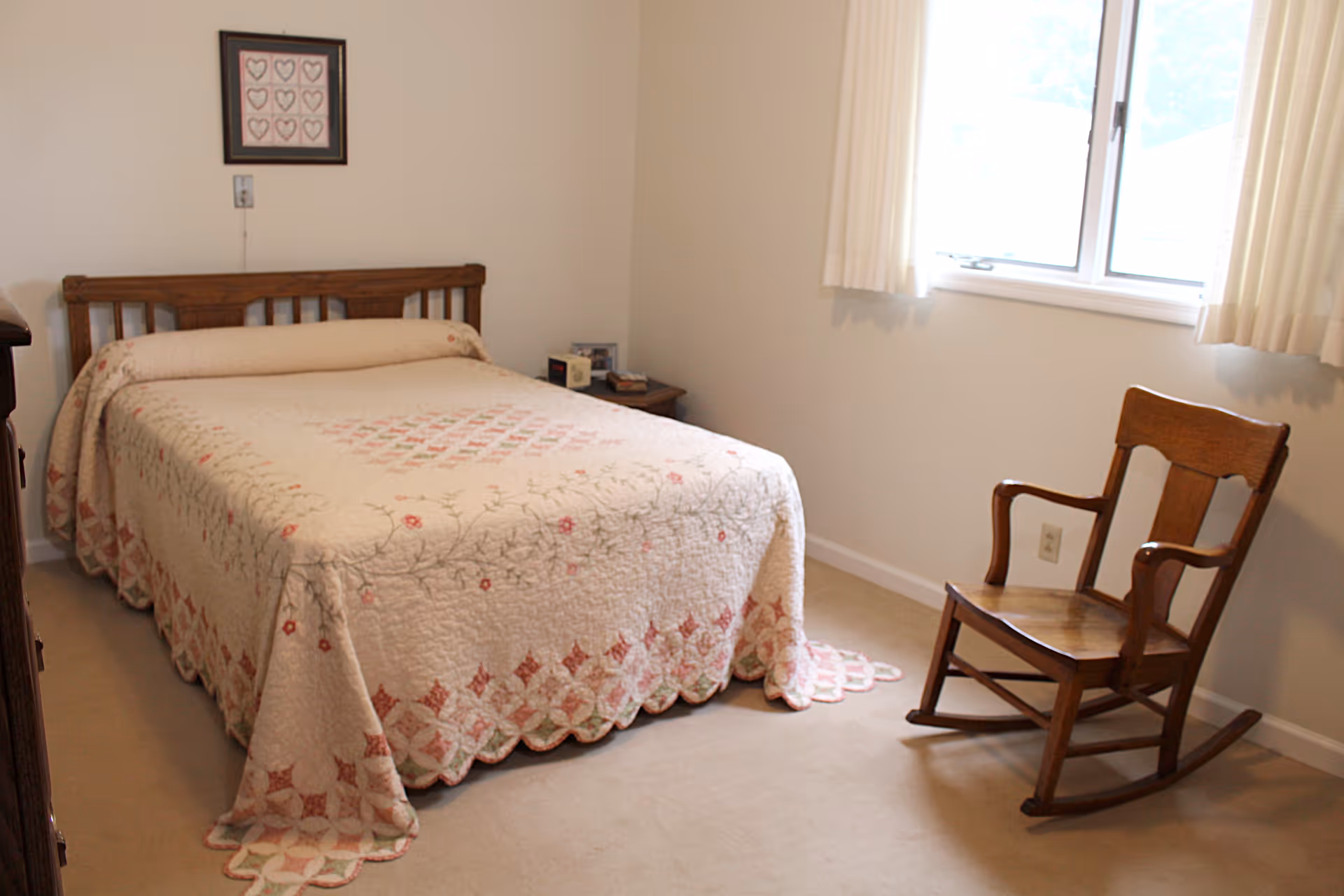 A simple bedroom with a wooden bed frame and a quilted bedspread featuring a floral pattern. There is a wooden rocking chair near a window with white curtains, and a small nightstand with a few items on it. A framed picture with heart designs hangs on the wall above the bed.