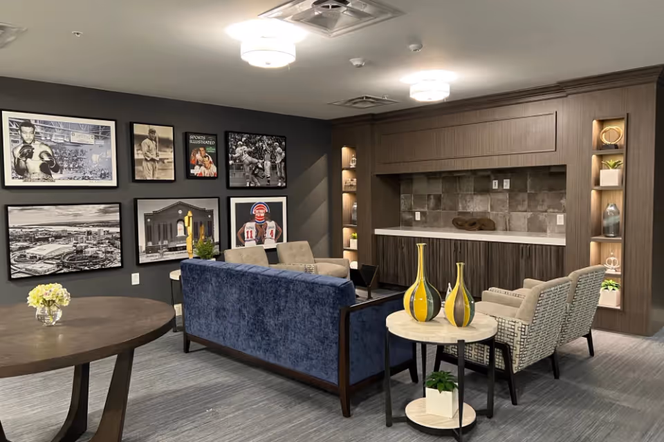 A modern lounge area with a blue velvet sofa, two patterned armchairs, and a small round table with decorative vases. The back wall features built-in wooden cabinetry with shelves displaying plants and decor items. The adjacent wall is decorated with framed black and white and color photographs, including sports and city scenes. The room has a gray carpet and ceiling lights.