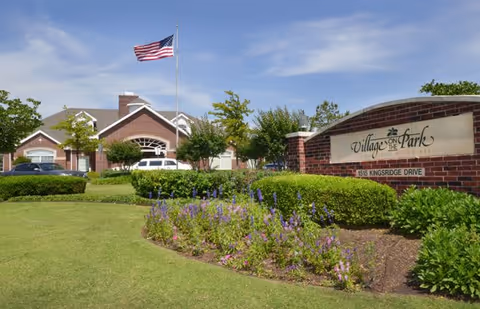 Front entrance of Village on the Park Oklahoma City senior living facility with a brick sign, lawn and flowerbeds, and an American flag flying in front.