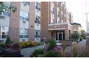 Exterior view of a multi-story brick building with large windows, featuring a landscaped garden area with colorful flowers and greenery in front of the entrance to Generations at Regency.